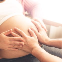 Pregnant couple of husband and wife feels love and relax at home. Young expecting woman holds baby in pregnant belly. Father take care of pregnant mother. Concept of maternity and pregnancy care.